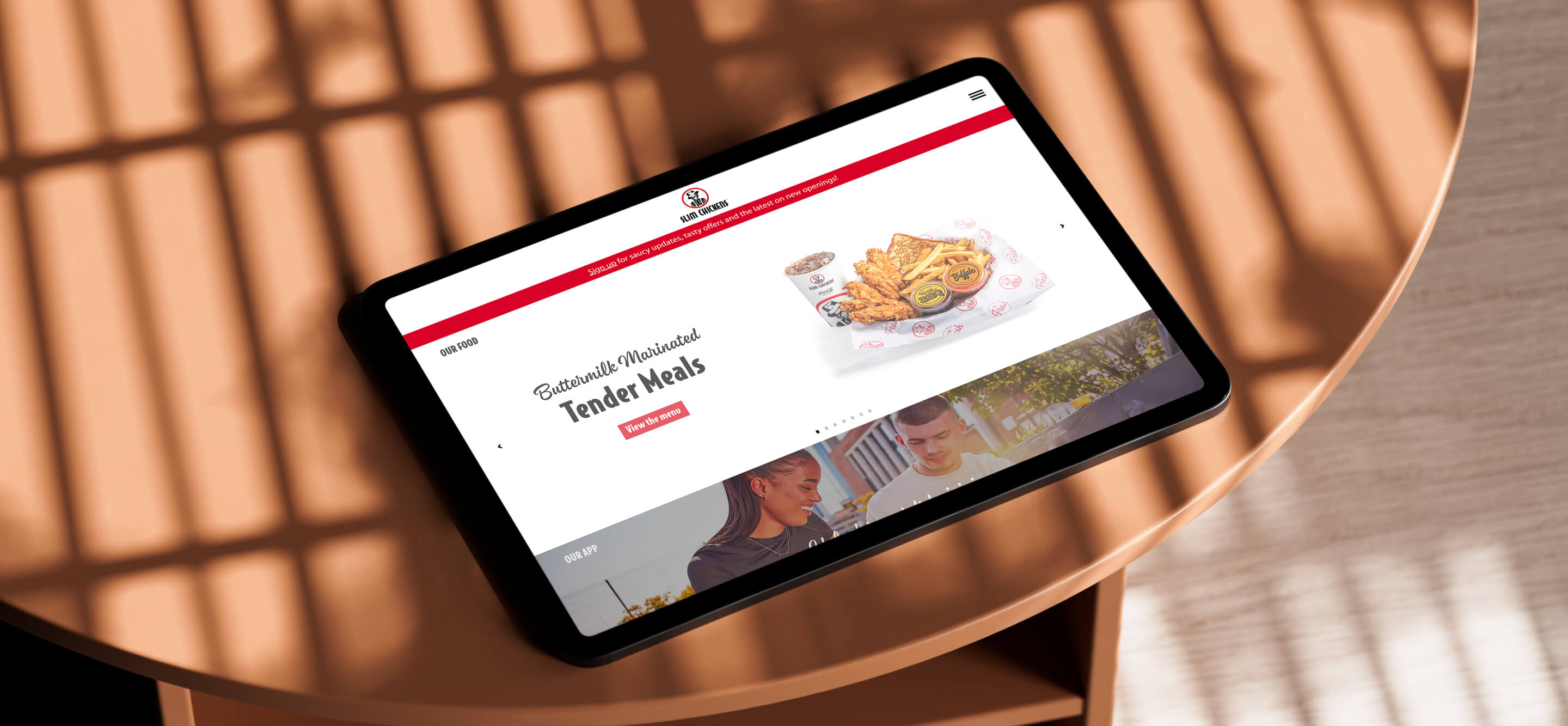 An image of the Slim Chickens home page on a tablet that is lying on a wooden table.